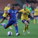 A player in a blue jersey tries to keep the ball as a player in a yellow jersey approaches her on a soccer field