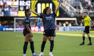 Trinity Rodman holds her arms in the air in celebration. Croix Bethune stands beside her. Both are smiling.